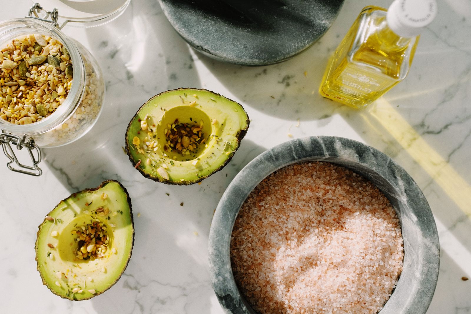 Avocado halves, olive oil, seeds and salt on marble — the healthy fats we were told to fear